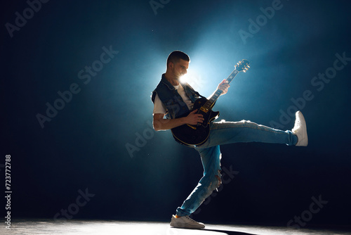 Casual rock artist on stage, with spotlights on stage behind him. Rock star playing intense solo against dark background with smoke. Concept of Rock-n-roll, music and dance, festivals and concerts.