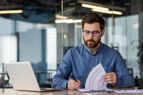 A young serious business man is sitting in the office at a desk with a laptop and is focused on working with documents. Writes and checks data and invoices