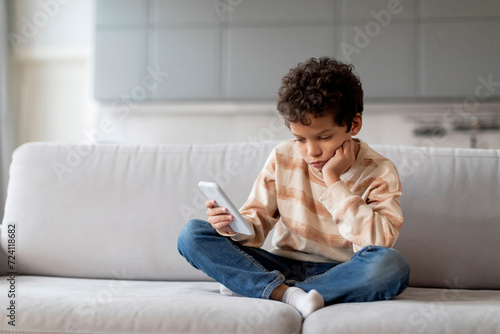 Little black boy sitting on sofa, looking disinterestedly at his smartphone