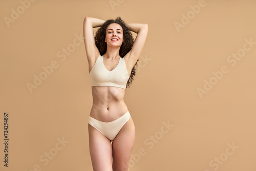 Hair removal. Confident slim woman wearing skin color bikini holding hands behind head and smiling at camera. Attractive female with wavy locks showing shaved smooth armpits over beige background.