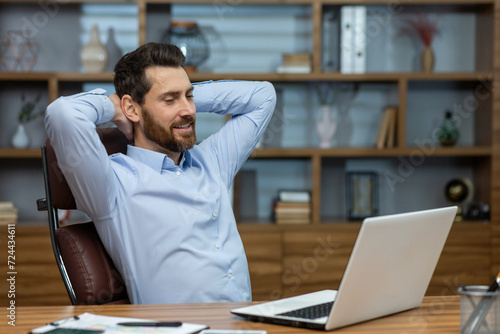 Relaxed brunette man leaning back and holding hands behind head in personal workspace with pc. Calm head of company taking break during busy day and enjoying daydreaming in comfortable chair.