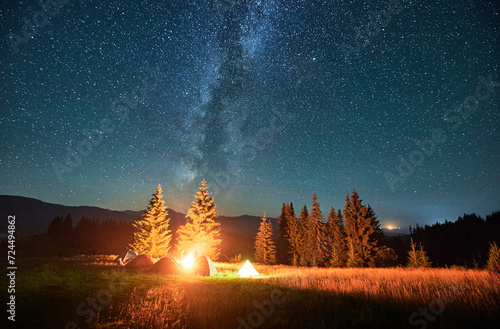 Night camping in mountains under starry sky and Milky way. Tents standing in campsite, fire burning, smoke raising to sky, stars shining. Concept of hiking in mountains.