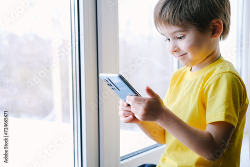Little toddler boy is holding the smartphone at home. He is wearing bright yellow t-shirt. Playing video games like a social problem