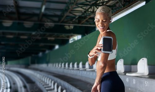 sport female athlete during running training and jogging, smiling African American woman touch a smartphone to armband, wear earphones and fitness smartwatch. Utilizing empty stadium stands and stairs