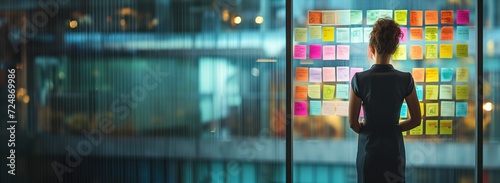 professional woman stares at a glass wall covered with colorful sticky notes. A planning concept, brainstorming or agile methodology in a business or creative environment.
