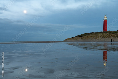 Low angle view of beach at low tide in front of historical red lighthouse and rising moon at on the island of Texel, the Netherlands during sunset and lighthouse and moon reflected in water pools