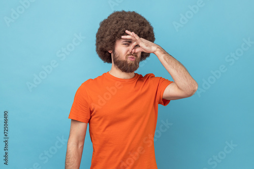 Portrait of man with Afro hairstyle wearing orange T-shirt looking at camera and nose in disgust, avoiding unpleasant smell of farting, stinky aroma. Indoor studio shot isolated on blue background.