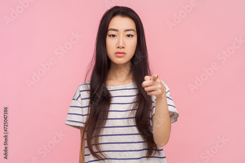 Portrait of bossy strict woman with long brunette hair pointing at camera choosing you displeased expression, wearing striped T-shirt. Indoor studio shot isolated on pink background.