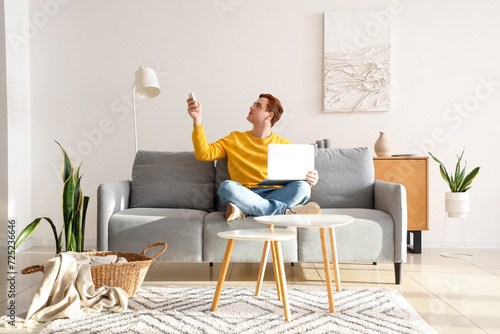 Young man with laptop turning on air conditioner on couch in living room