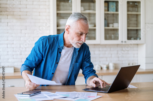 Mature man look on screen on laptop, working at home with documents.