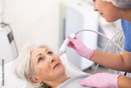 Pleased aged woman having vacuum cleaning of her face by means of apparatus for aesthetic procedures used by specialist