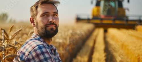 Contented farmer in a field, combine harvester operator approaching bountiful wheat harvest, agronomist in plaid shirt gazing at camera on a farm