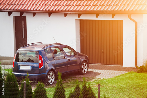 A blue family car sits peacefully in the driveway of a beautiful detached house with a brown garage door and red roof tiles.