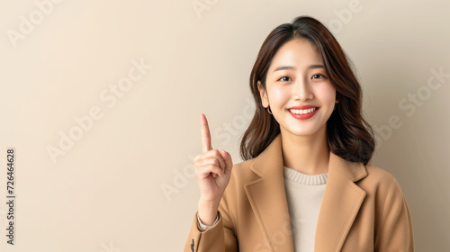 Portrait of a Young Korean woman in a beige coat gesture with the index finger pointing upwards isolated with copy space.