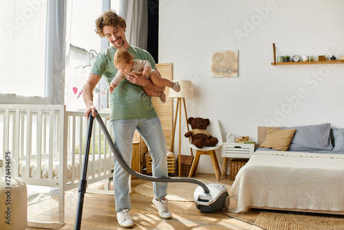 man multitasking housework and childcare, father vacuuming hardwood floor with infant boy in arms