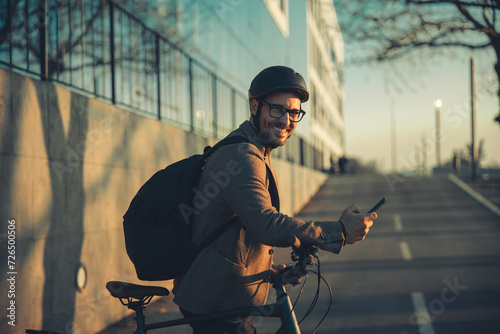 A portrait of a happy businessman with a backpack using a smartphone and leaning on handlebars of a bicycle
