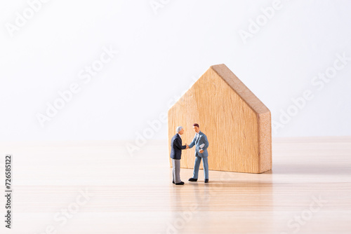 木製の住宅模型の前で握手するミニチュアビジネスマン