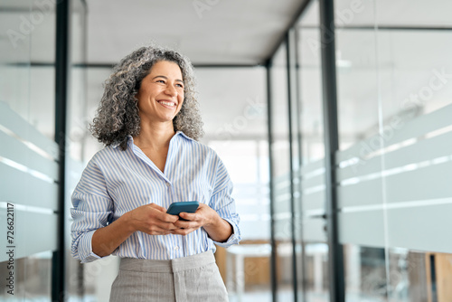 Happy middle aged business woman holding mobile cell phone using cellphone in office. Smiling mature old professional lady business investor owner entrepreneur using smartphone standing looking away.