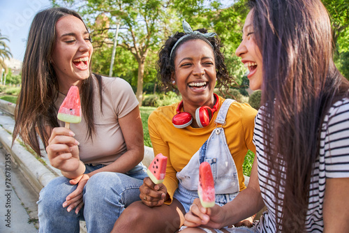 Group of young multiracial female companions laughing enjoying ice cream in park on summer day. Three funny women smile excitedly while chatting outdoors. Generation z and friendship in youth.