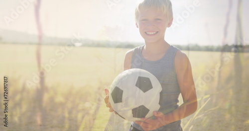 Image of plants and sunlight over happy caucasian schoolboy with football in field