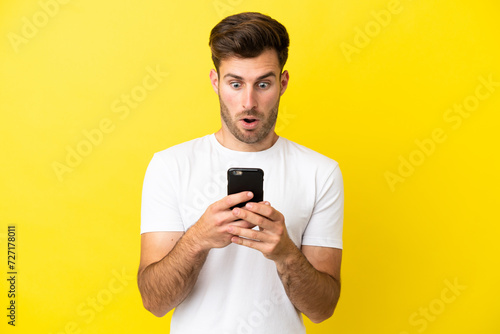 Young caucasian handsome man isolated on yellow background looking at the camera while using the mobile with surprised expression