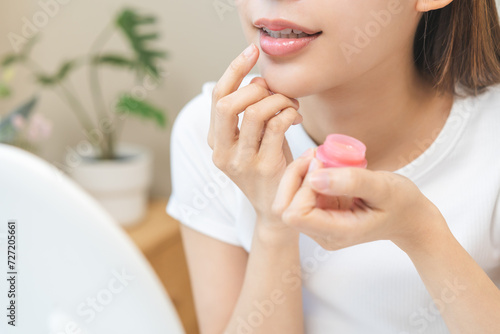 Lip care concept, happy asian young woman finger touching lips in front of mirror at home after applying lipstick balm on dry mouth from natural beauty product, skincare routine, makeup and cosmetics.