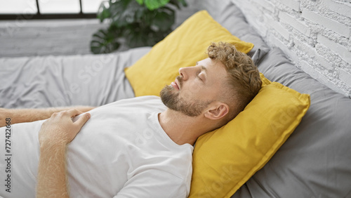 Young bearded man sleeping peacefully in a modern bedroom with yellow pillows and gray bedding.