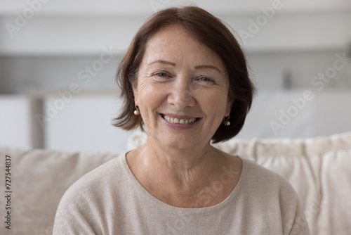 Positive mature older retired woman home head shot portrait. Female pensioner sitting on sofa, looking at camera with happy smile, enjoying retirement, talking on video conference call