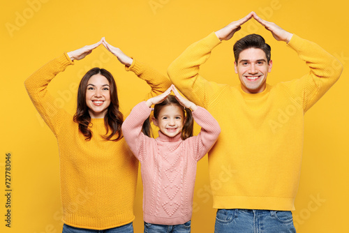 Young parents mom dad with child kid girl 7-8 year old wear pink knitted sweater casual clothes holding folded hands above head like house roof isolated on plain yellow background. Family day concept.