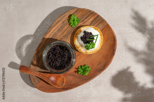 Black caviar on mini blini or pancake with glass jar on a wooden plate.