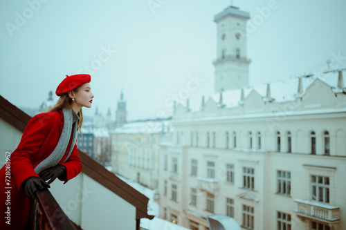 Fashionable woman wearing red beret, elegant winter coat, knitted gray scarf, gloves, posing on balcony with beautiful view on snow covered European city Lviv. Copy, empty, blank space for text