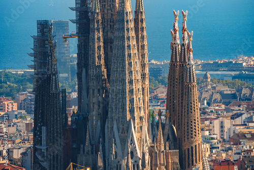 Closeup of Sagrada Familia's spires in Barcelona, showcasing Gaudi's Gothic and Art Nouveau design, with cranes indicating ongoing construction against a cityscape backdrop.