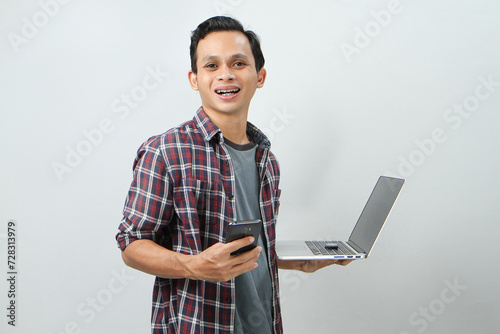 Happy asian indonesian man holding smartphone and laptop computer on isolated background