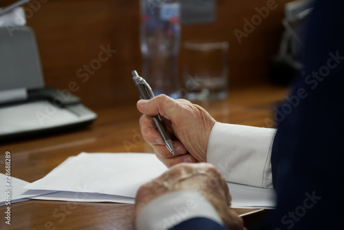 An elderly man in a business suit with cuffs, sitting at a desk with pen and documents. Concept of writing a will, drafting a contract or filling out forms. Photo. No face. Selective focus