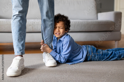 Upset child boy clinging to father's leg at home