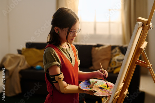 Pretty pre-teen girl with disability standing in front of easel and taking mixed watercolor from palette in myoelectric hand