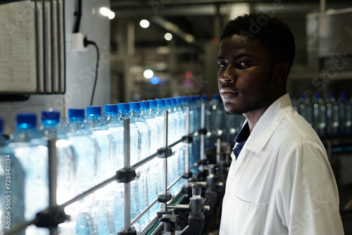Young African American quality control engineer in labcoat standing in front of assembly line with capped bottled drinks