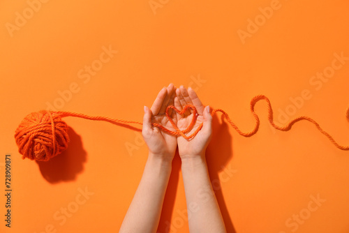 Female hands holding thread of yarn ball with heart on orange background