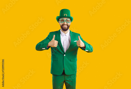 Happy bearded man enjoying cool great party on St Patrick's Day. Cheerful joyful man in green costume, hat and funny shamrock clover glasses smiles and gives thumbs up isolated on yellow background