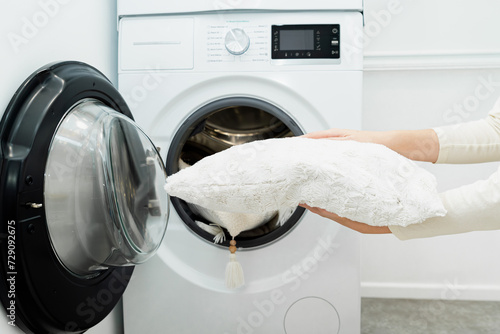 Woman holding clean white pillow in front of the drum of washing machine in laundry room. Washing pillows