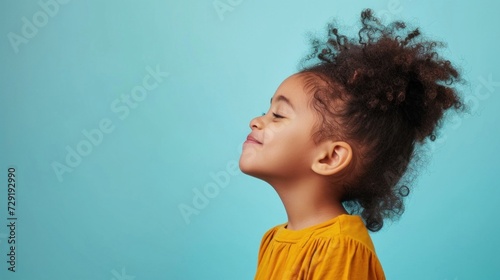 A young child with curly hair wearing a yellow top looking up with a smile against a light blue background.