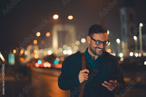 A satisfied mid adult businessman with glasses reading text messages on his phone and walking back home during night