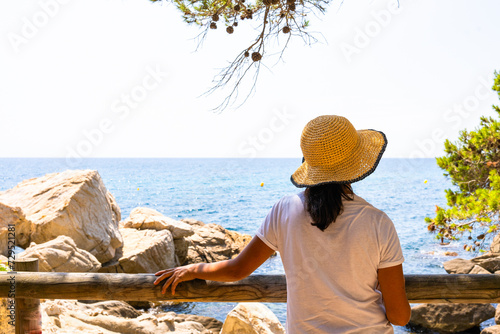 Woman on her back with a hat contemplating the horizon in a beautiful view towards the sea on the Spanish Costa Brava surrounded by pine trees and with intense summer light
