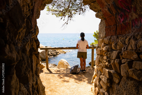 Unrecognizable woman from behind enjoying beautiful views of the Mediterreans sea from above and framed by a stone construction on a summer day