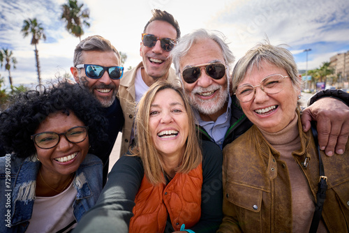 Group of diverse cheerful middle-aged tourist friends posing smiling taking a photo selfie embracing together with front camera on travel outdoor. Mature happy six people enjoy holidays sunny day 