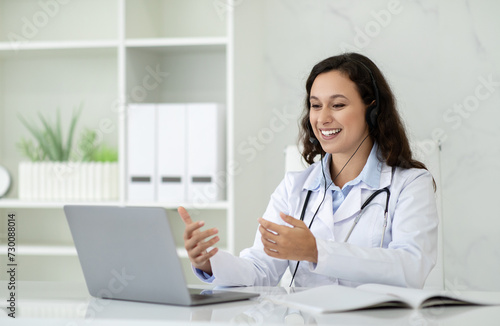 Cheerful young brunette lady in white coat has video call
