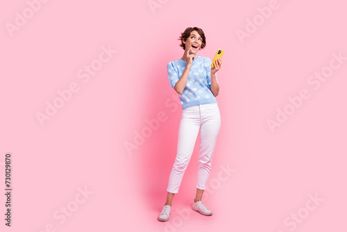 Full size portrait of pretty creative girl finger touch chin hold smart phone look empty space isolated on pink color background