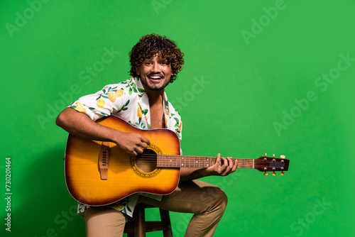 Photo portrait of attractive young man sit chair play guitar dressed stylish citrus print clothes isolated on green color background