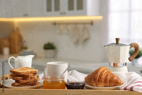 Breakfast served in kitchen. Toasts, honey, jam, fresh croissant, coffee and pitcher of milk in wooden tray, closeup