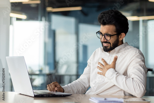 Overworked indian businessman feeling unwell at desk with laptop in modern office